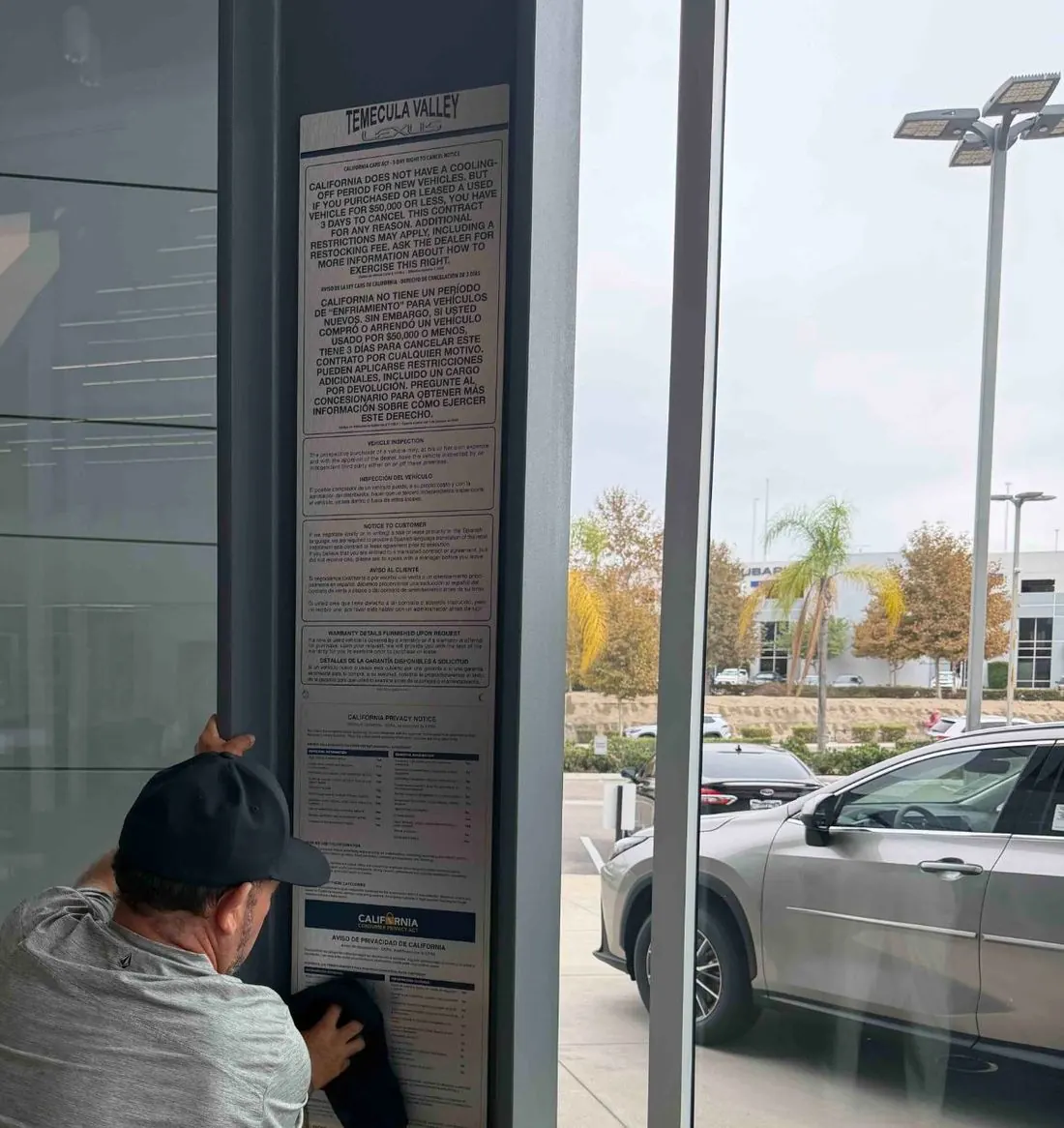 An installer finishing a brushed-aluminum combo disclosure board with the new CARS Act notice on a showroom pillar